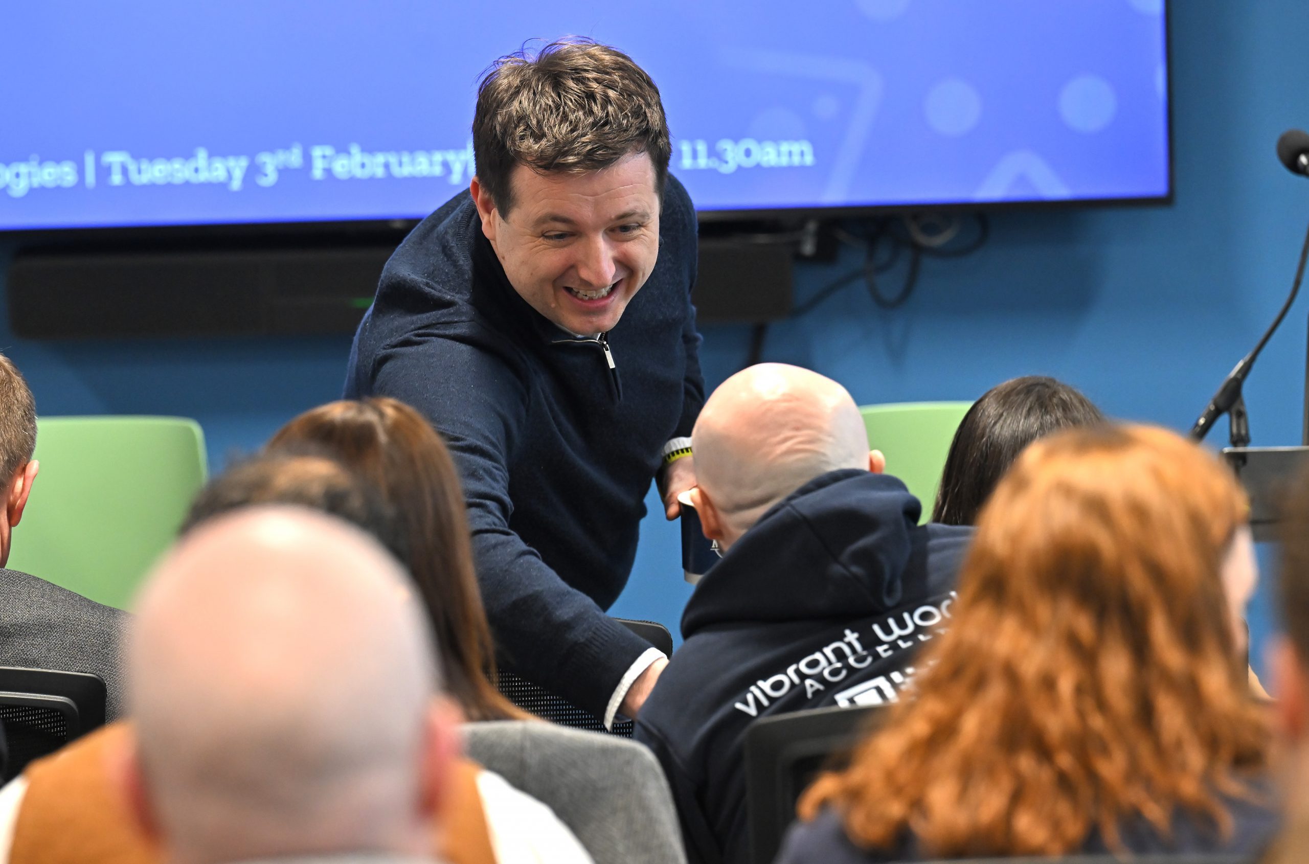 An attendee leans forward to speak to someone seated in the first row before a presentation begins.