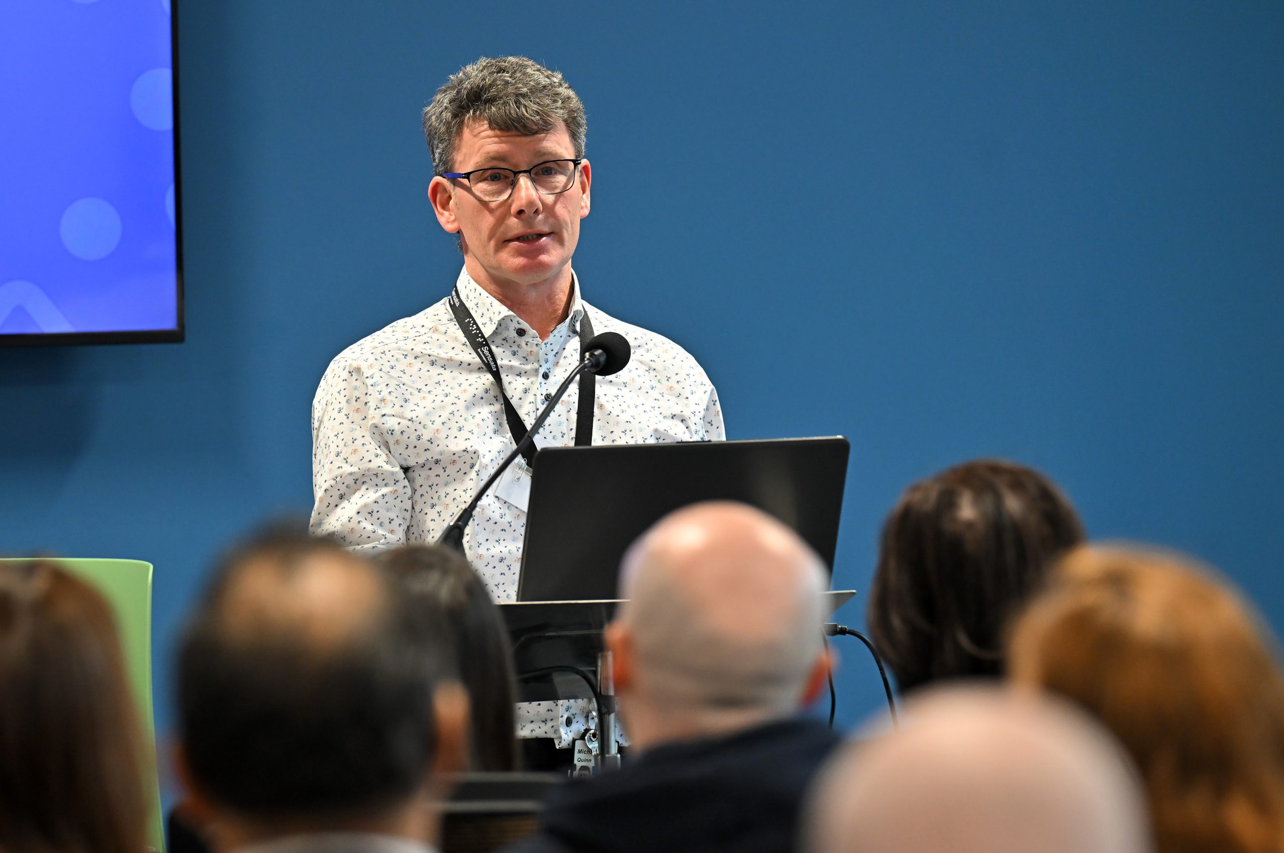 A speaker stands at a podium addressing an audience, with a microphone and laptop in front of him.