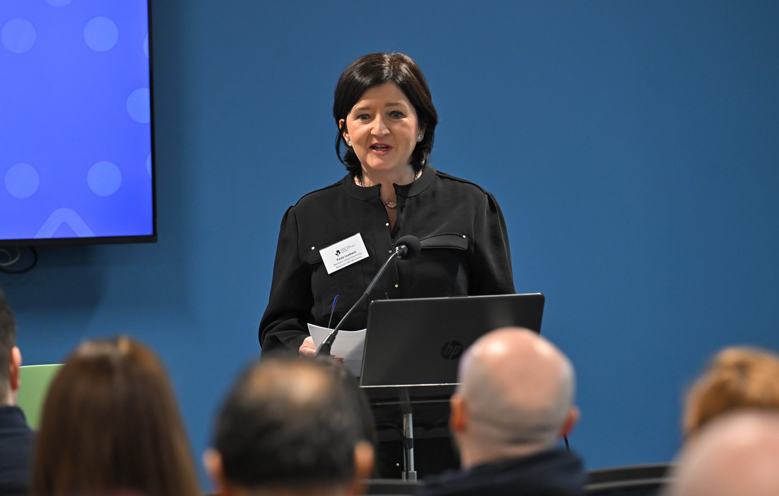 A speaker in a black outfit delivers a presentation from a podium while the audience listens.
