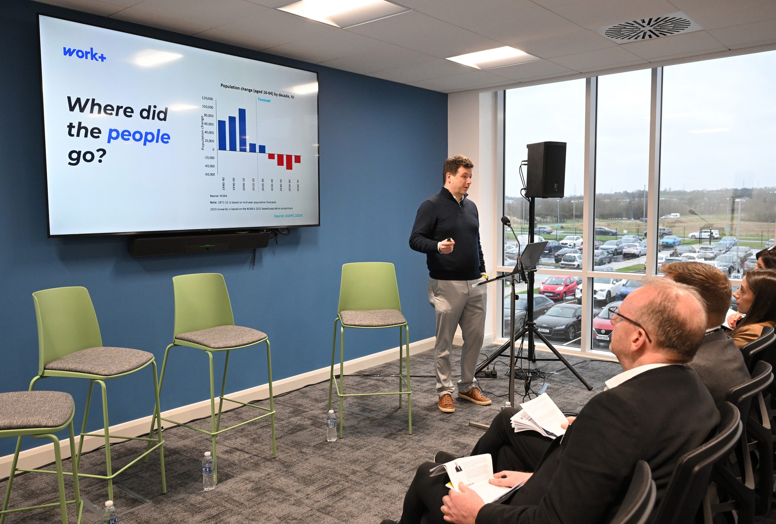 A presenter stands beside a large screen showing a chart titled “Where did the people go?” while the audience watches.