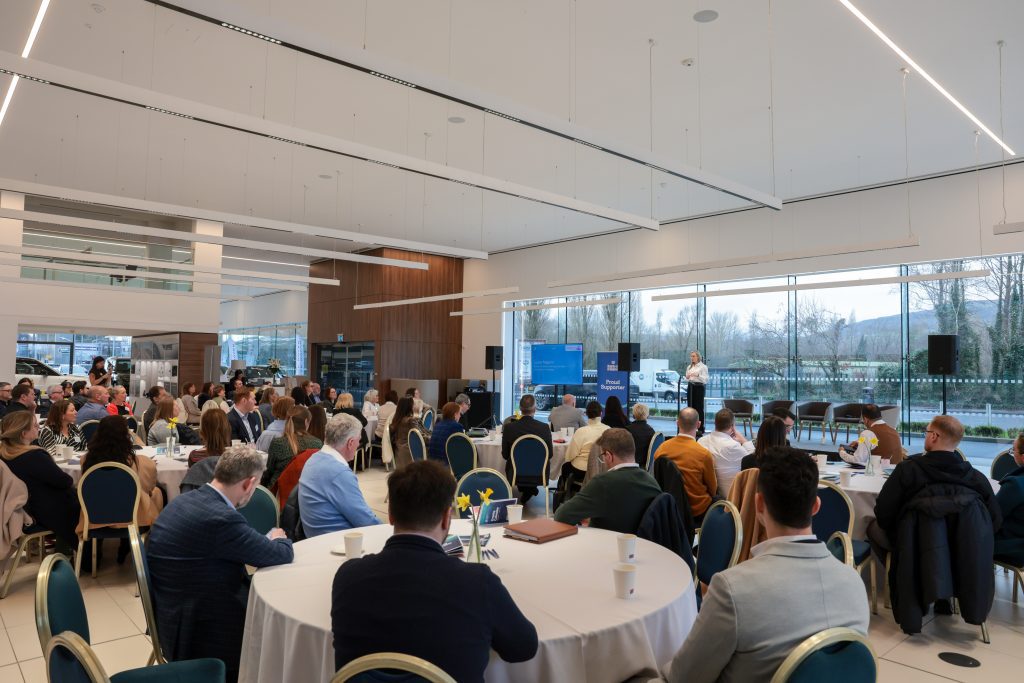 Attendees seated at round tables listening to a keynote speaker at the front of a modern venue.