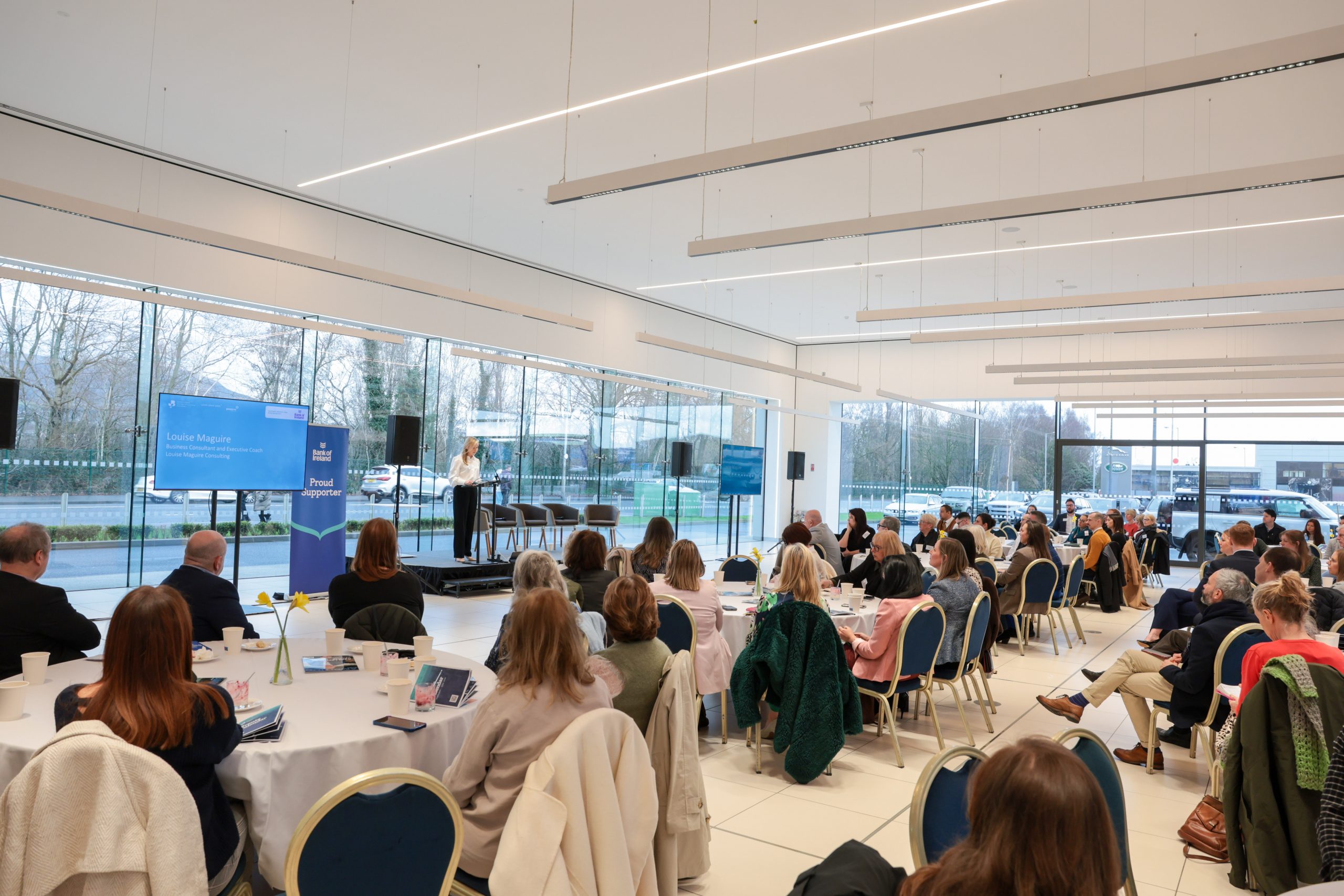 Attendees seated at round tables listening to a keynote speaker presenting at the front of a bright, modern event space with large windows.