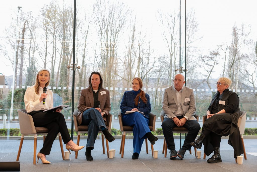 A panel of five speakers seated on stage, participating in a discussion in front of large windows.