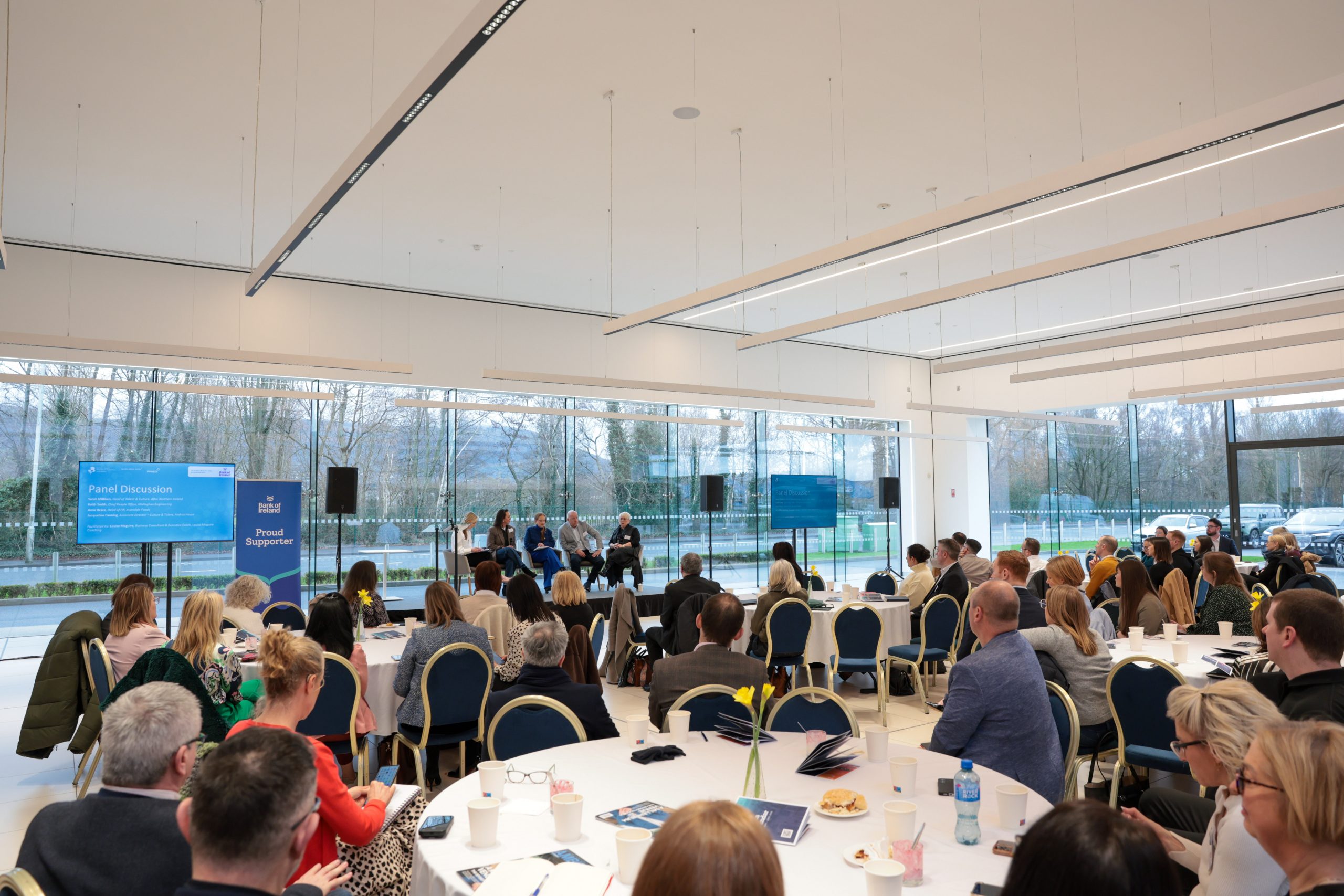 A large conference room with round tables filled with attendees listening to a panel discussion on a stage, with floor‑to‑ceiling windows in the background.