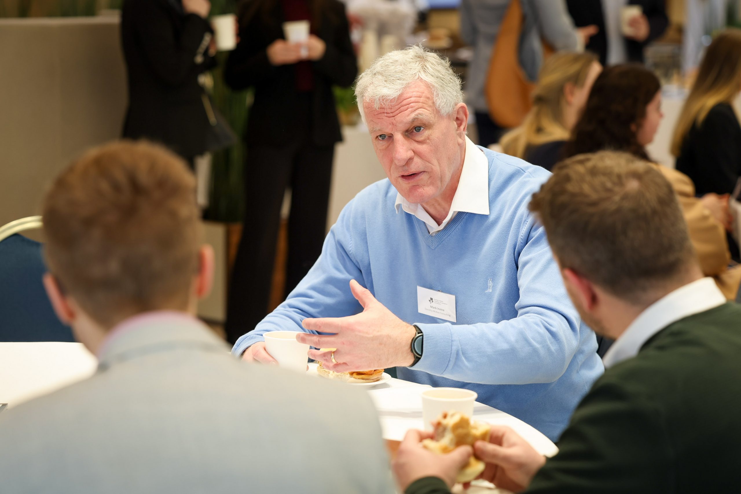An attendee in a light blue top speaking to two others at a table during a networking break.