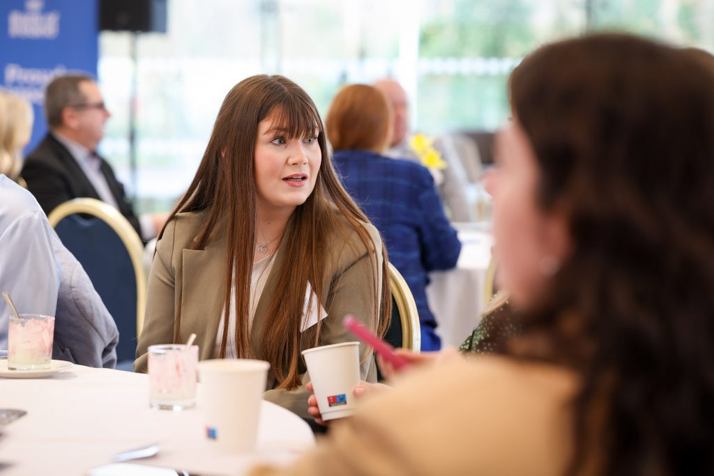 A person seated at a table holding a drink cup while listening to someone speaking nearby.