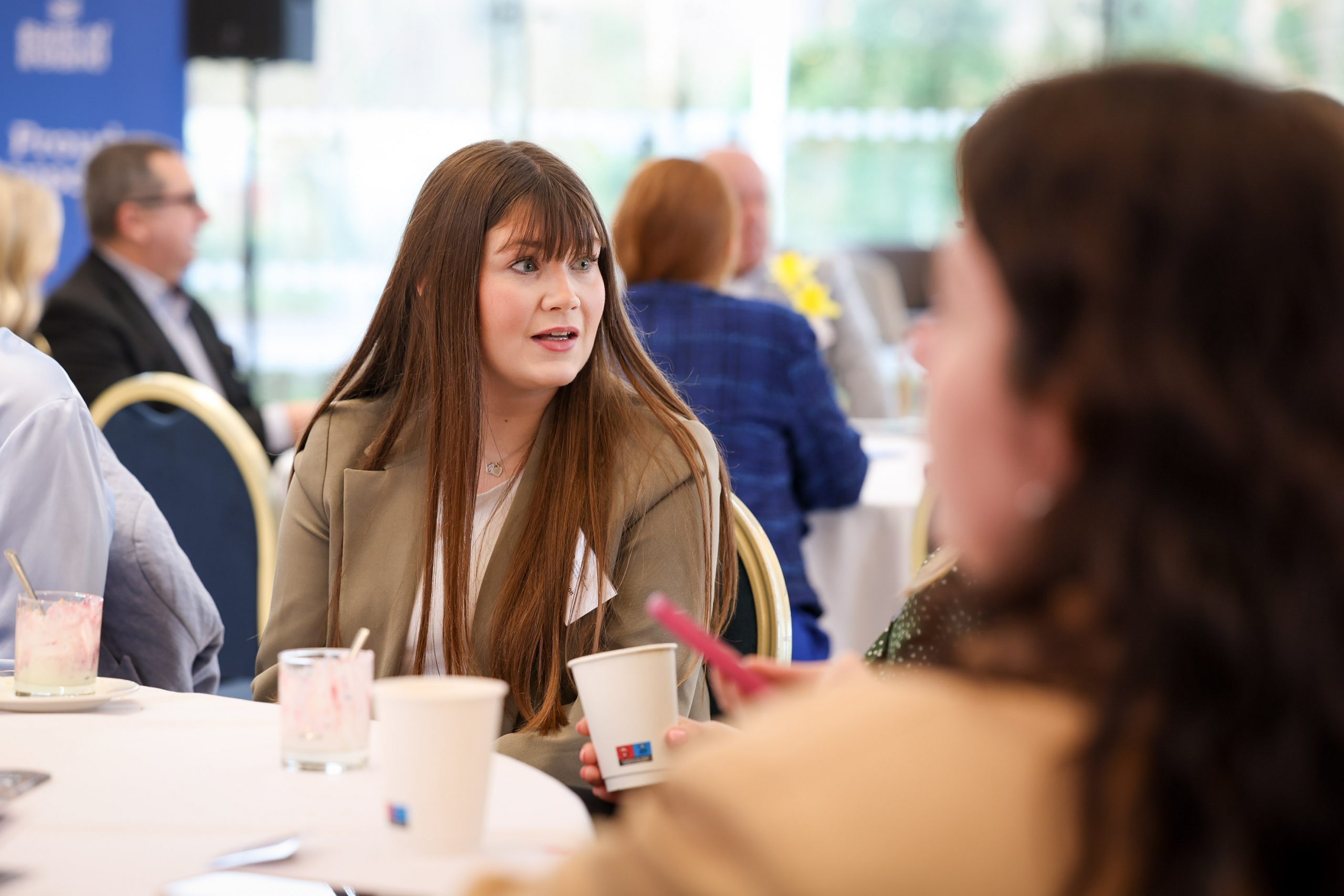 A person seated at a table holding a drink cup while listening to someone speaking nearby.