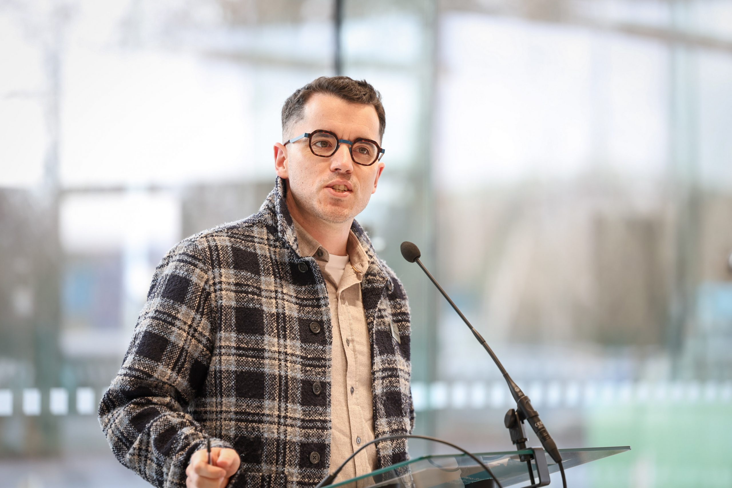 A speaker addressing the audience from behind a lectern, with a microphone and glass podium visible.