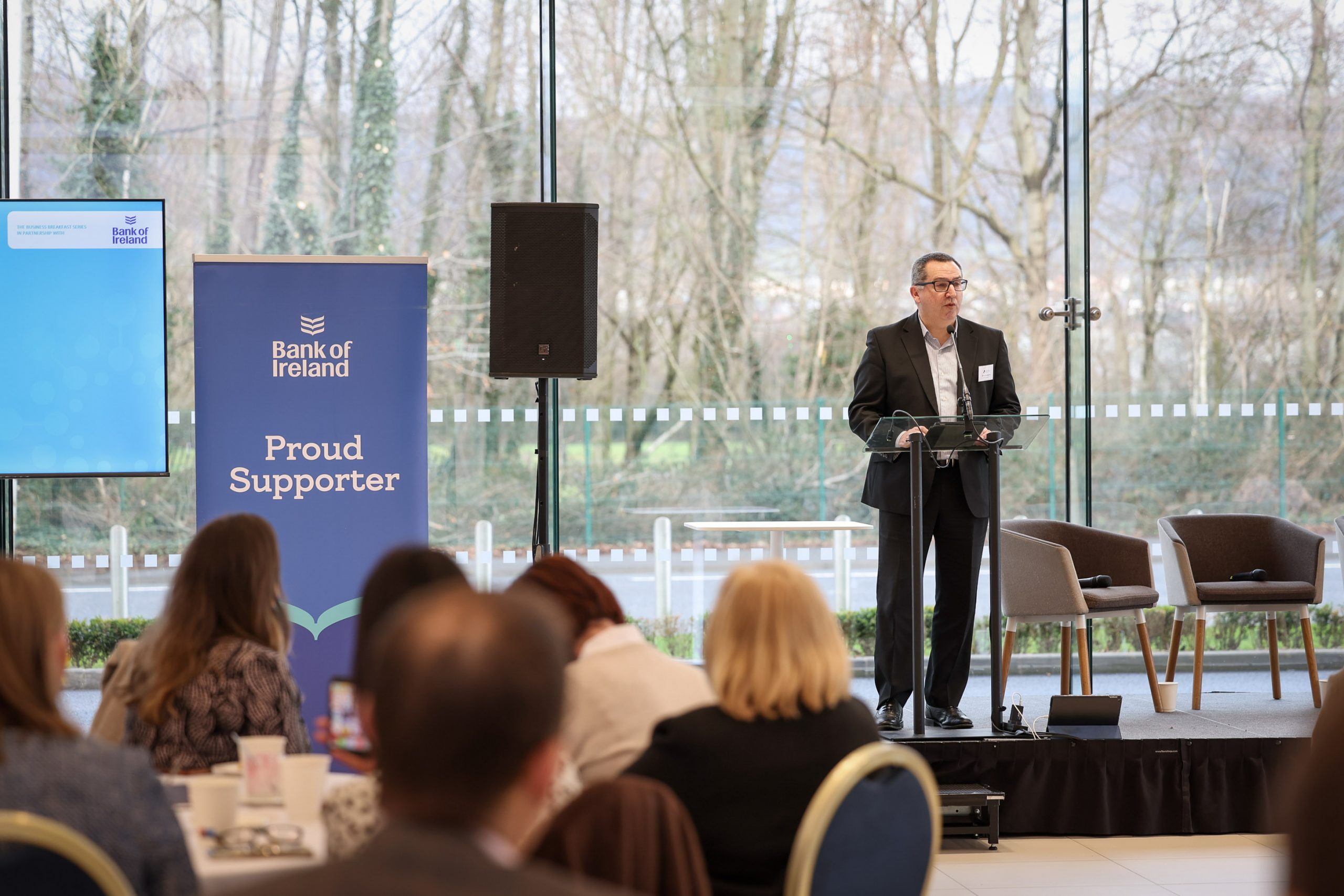 A speaker presenting at a lectern during an event, with branding and screens visible behind them.