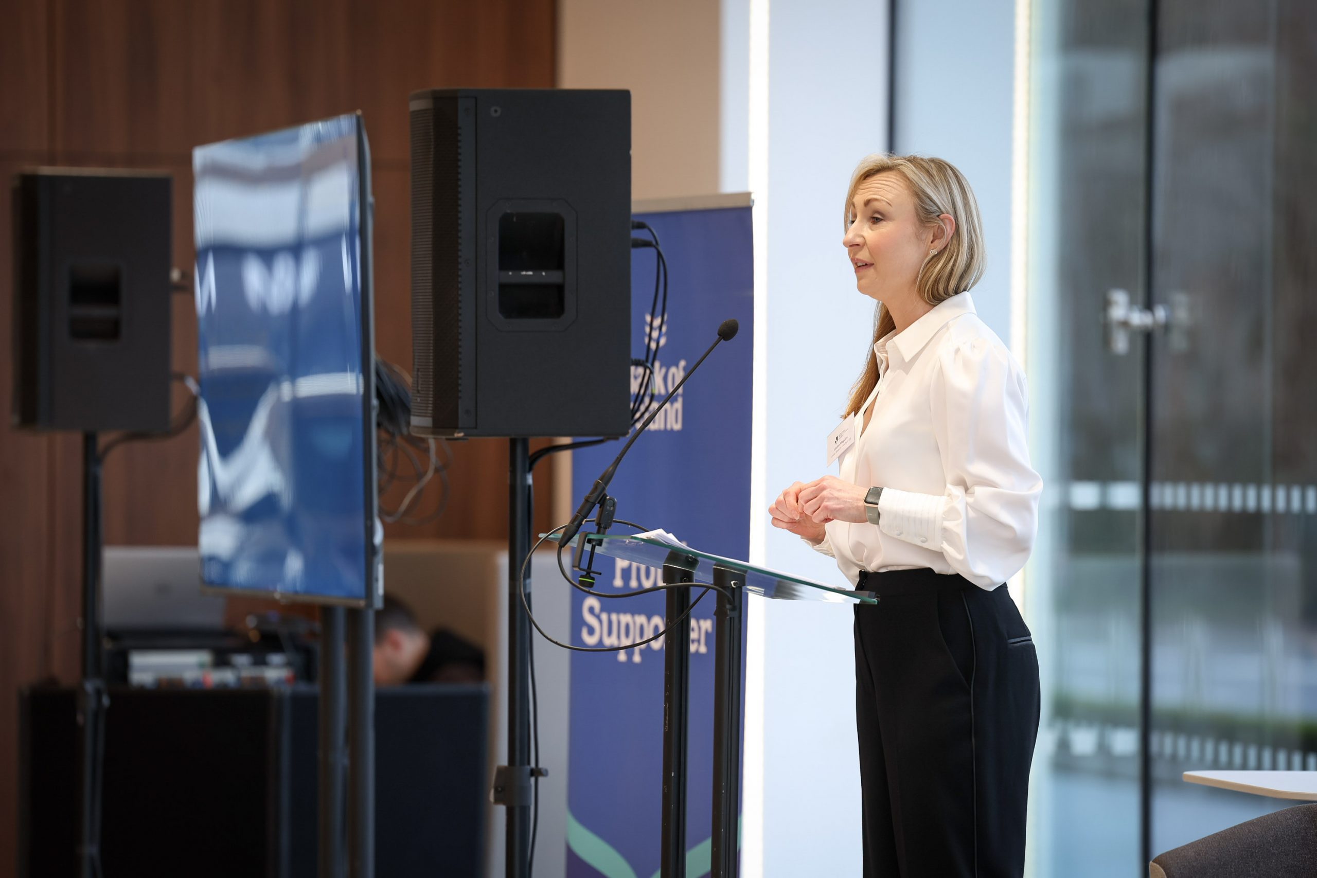 A speaker standing at a podium presenting to an audience, with event branding displayed beside them