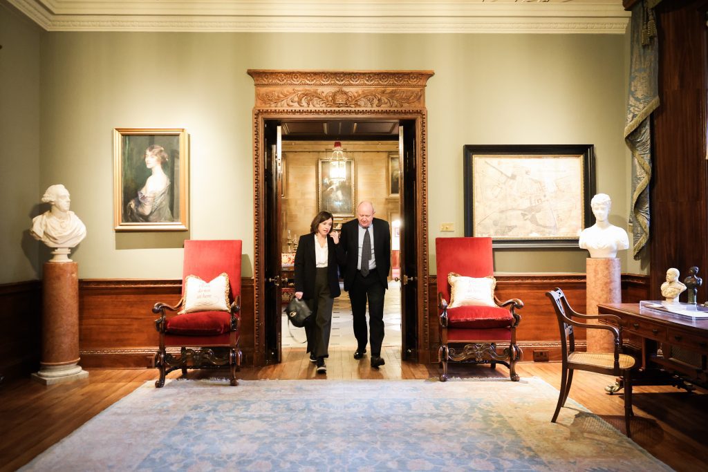 Two people walking and talking inside an ornate room featuring red chairs, bust sculptures, wood panelling, and framed artwork.