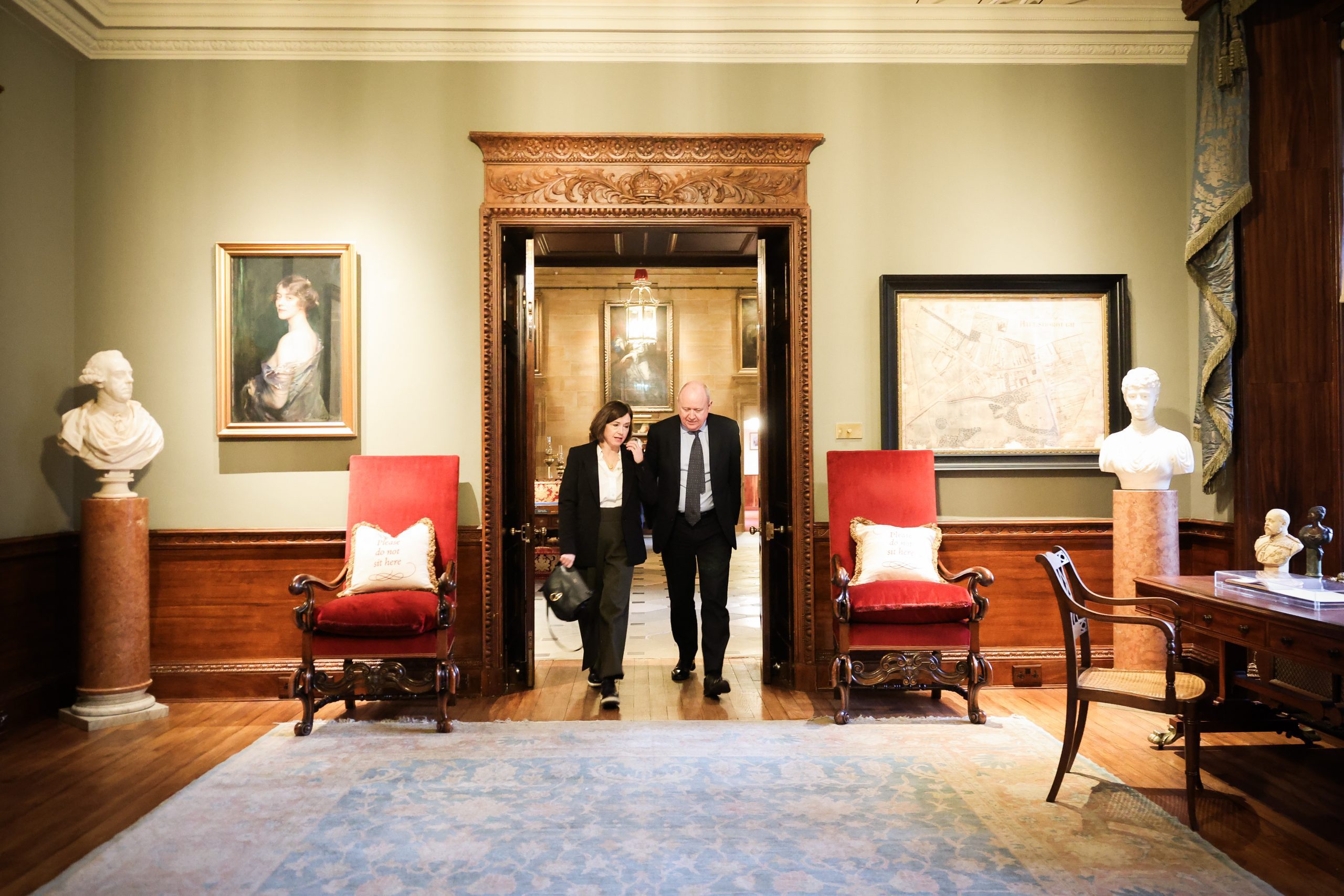 Two people walking and talking inside an ornate room featuring red chairs, bust sculptures, wood panelling, and framed artwork.
