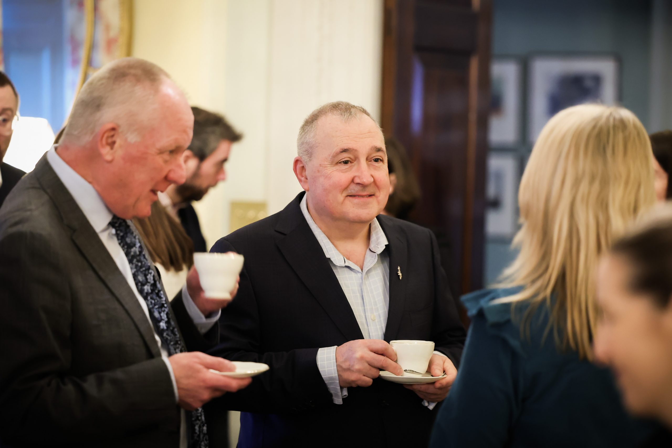 Several people gathered indoors at an event, holding cups and saucers, with framed artwork and a doorway in the background.