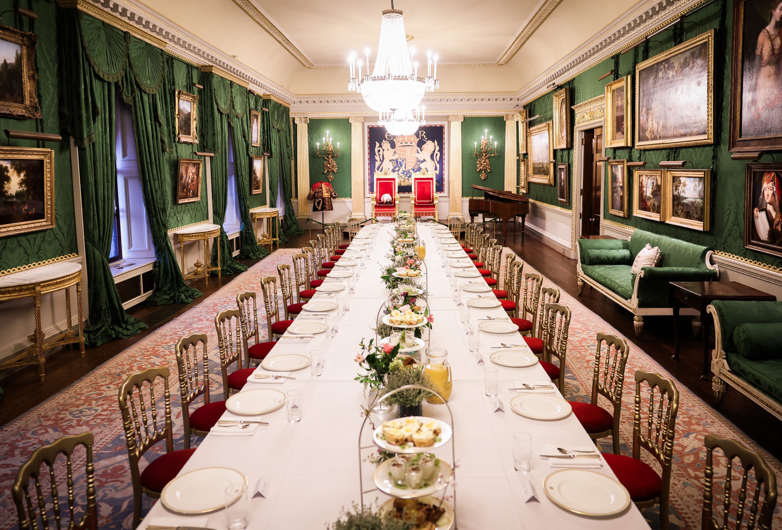 A long, formal dining room decorated with green drapes, chandeliers, and framed artwork, with a table fully set for an event.