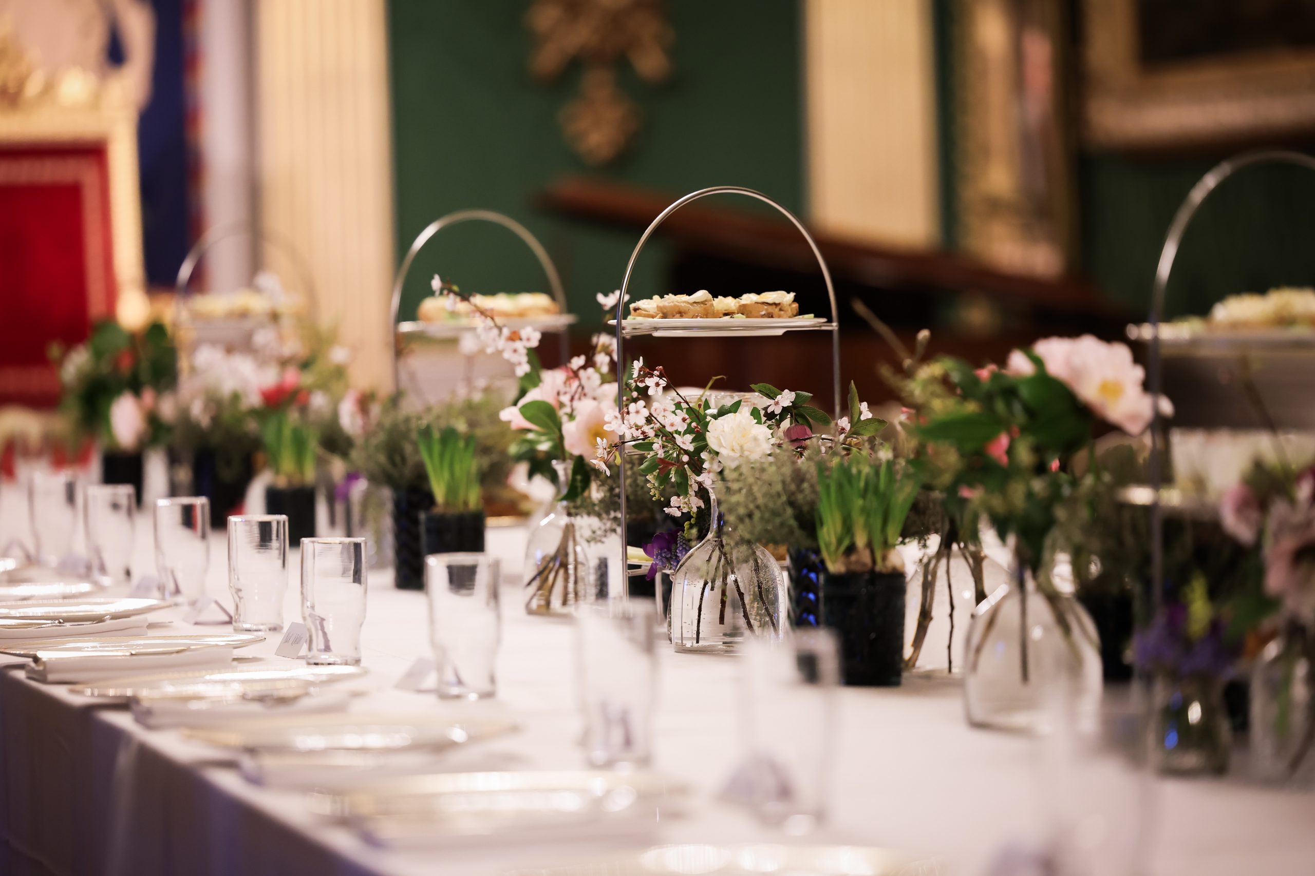 A long dining table set with glassware, plates, floral arrangements, and tiered trays of food in a formal room.