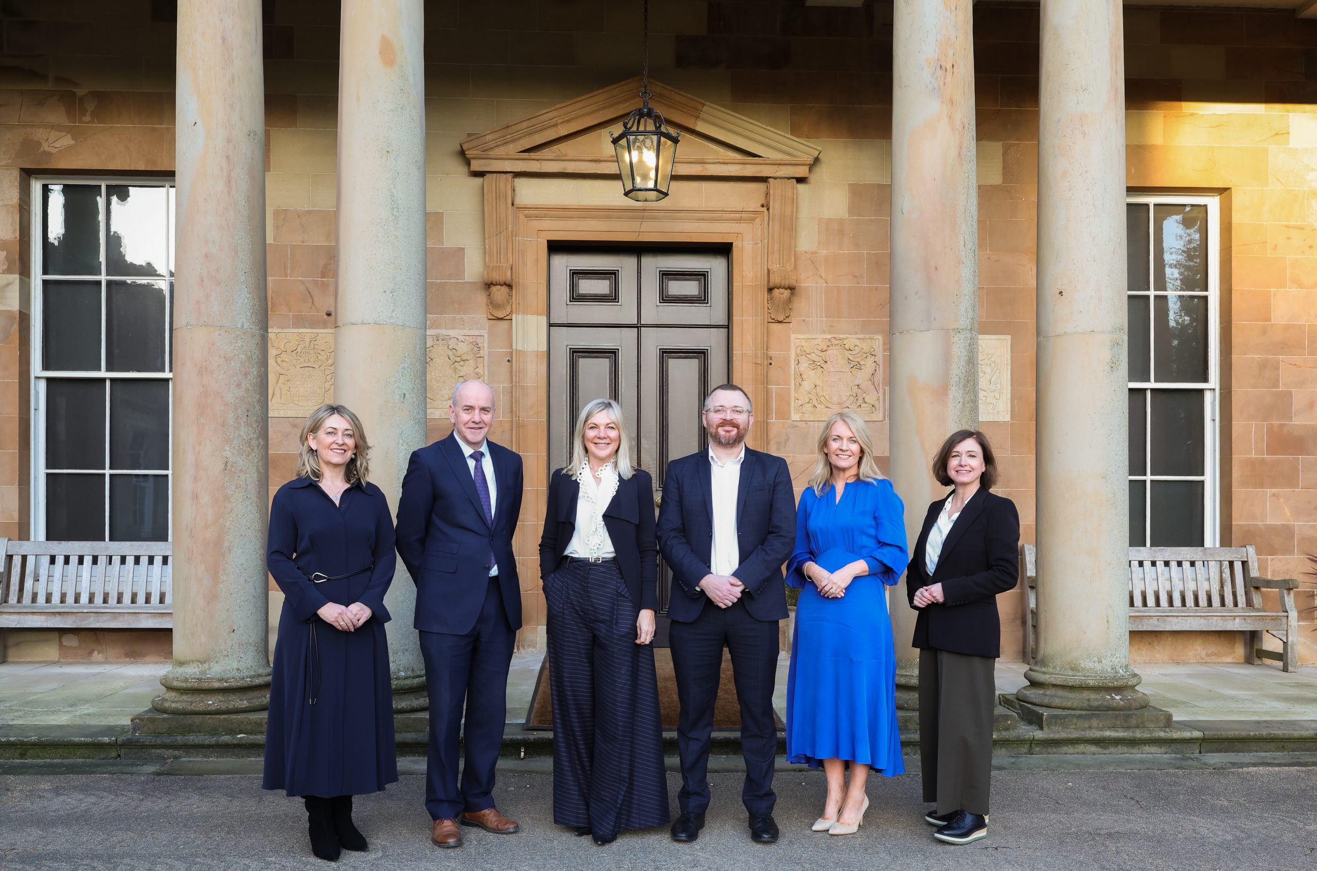 A group of six people standing in front of a columned entrance to a historic building, posed for a group photo.
