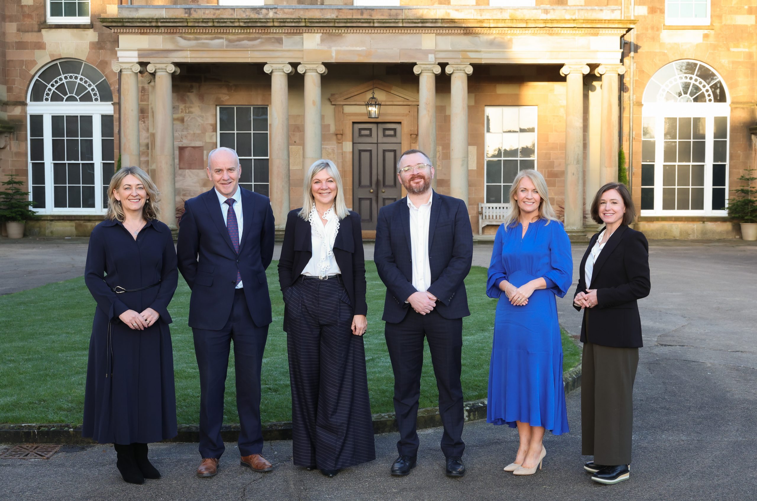 Six people standing outdoors in front of a large building with tall windows and columns, arranged side by side for a group photo.