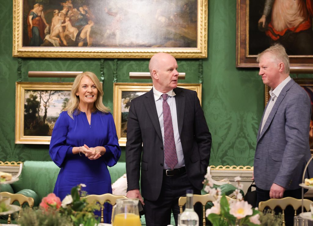 Three people standing and talking in front of a dining table, with several framed paintings displayed on green walls behind them.