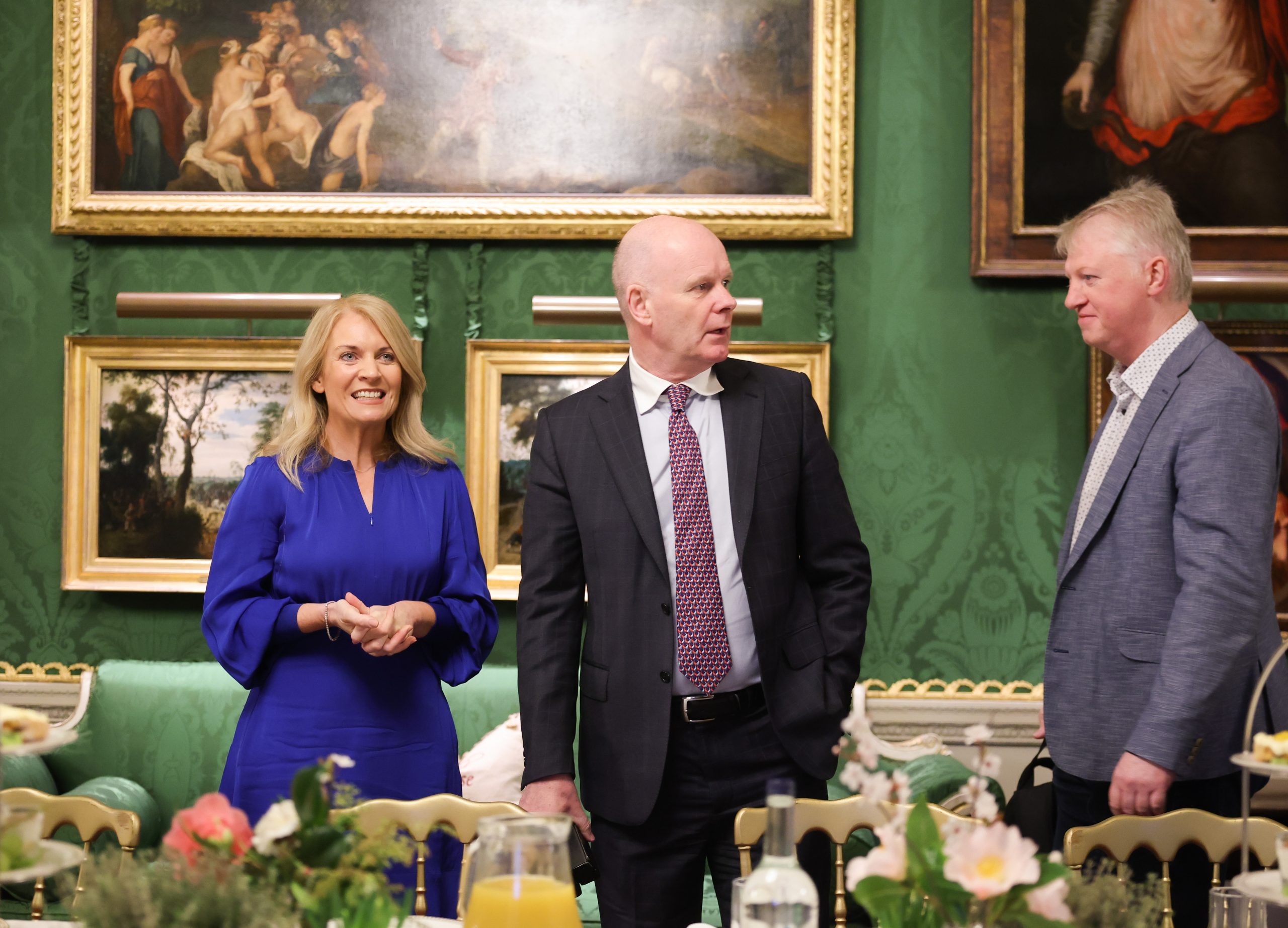 Three people standing and talking in front of a dining table, with several framed paintings displayed on green walls behind them.