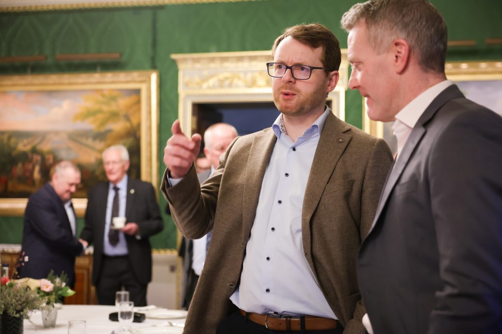A person gesturing while speaking to another attendee during a gathering in an ornate room with green walls and framed paintings.