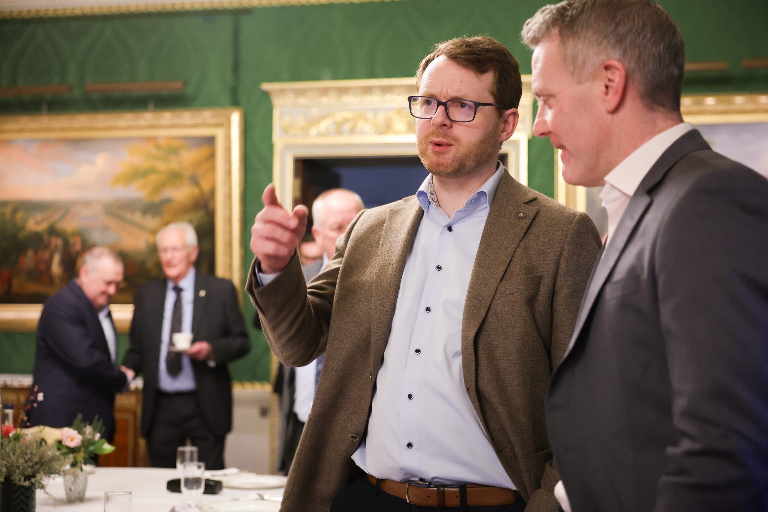 A person gesturing while speaking to another attendee during a gathering in an ornate room with green walls and framed paintings.