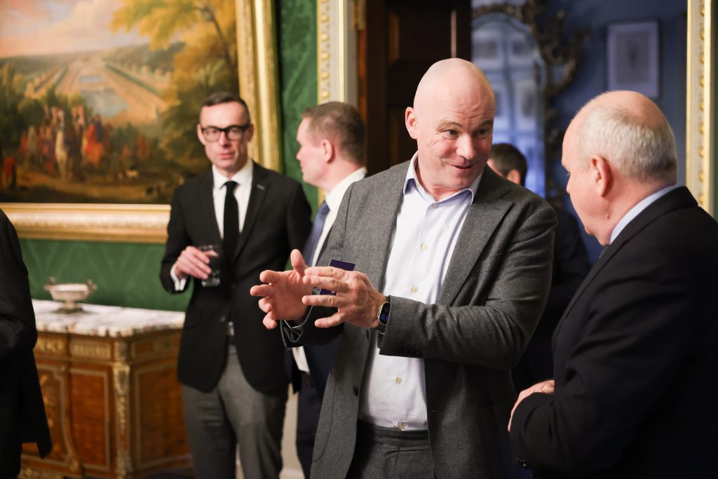 Several people in conversation during a formal event, with one person gesturing while others listen, surrounded by ornate decor and a large wall painting.