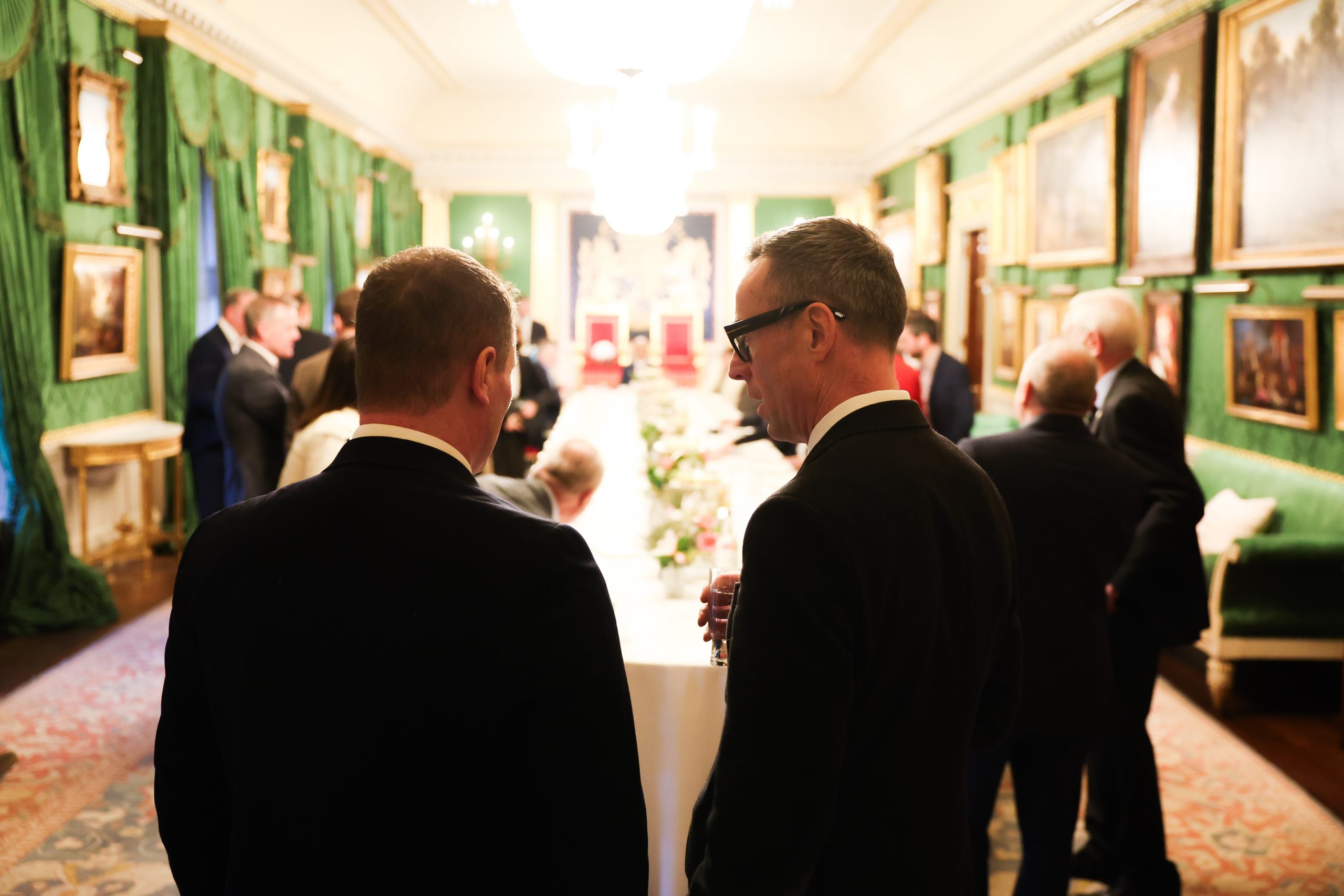 Two people standing and talking near a long dining table in an ornate room, with several others gathered in the background.