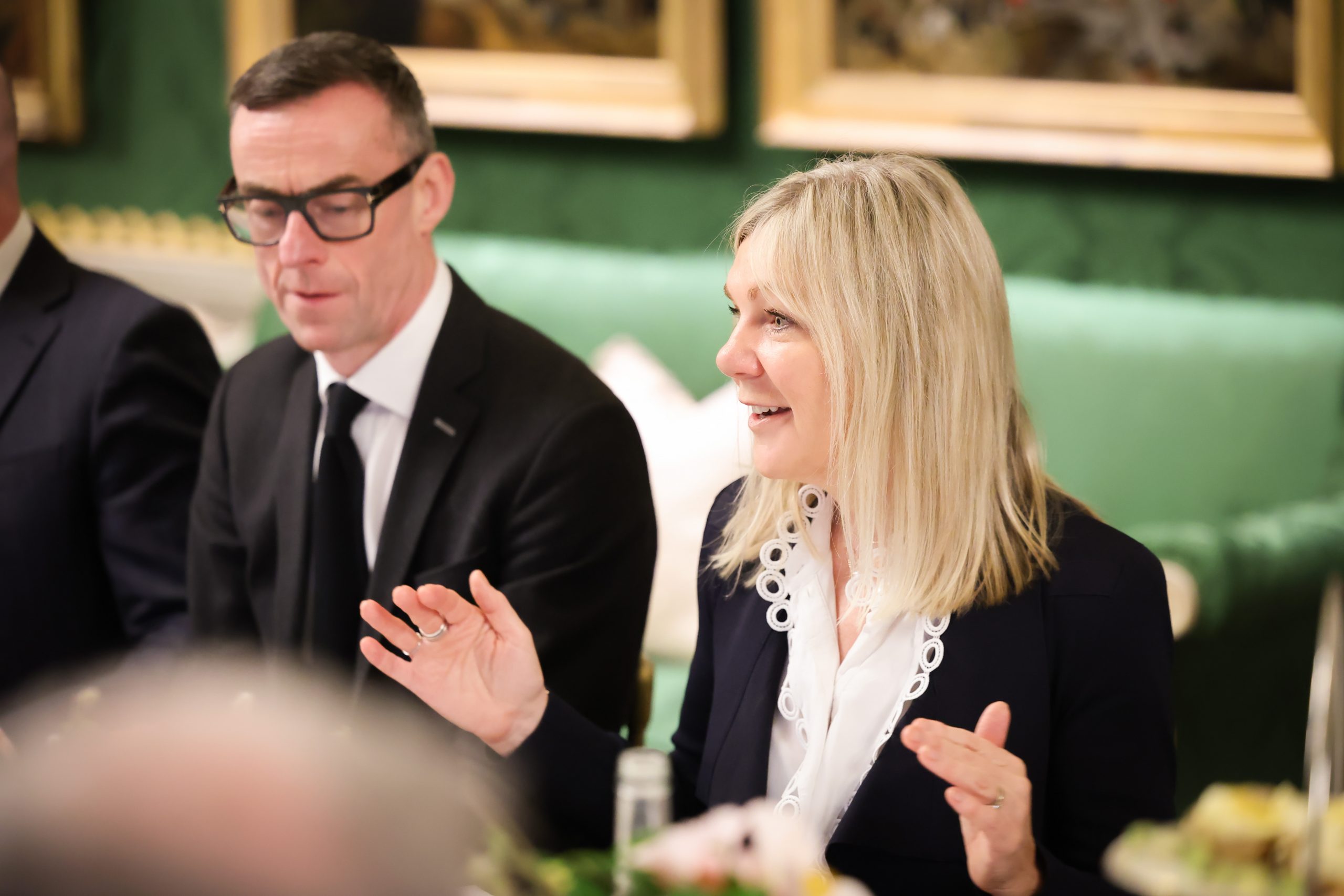 Two people seated at a formal table engaged in conversation, with decorative green walls and framed artwork in the background.