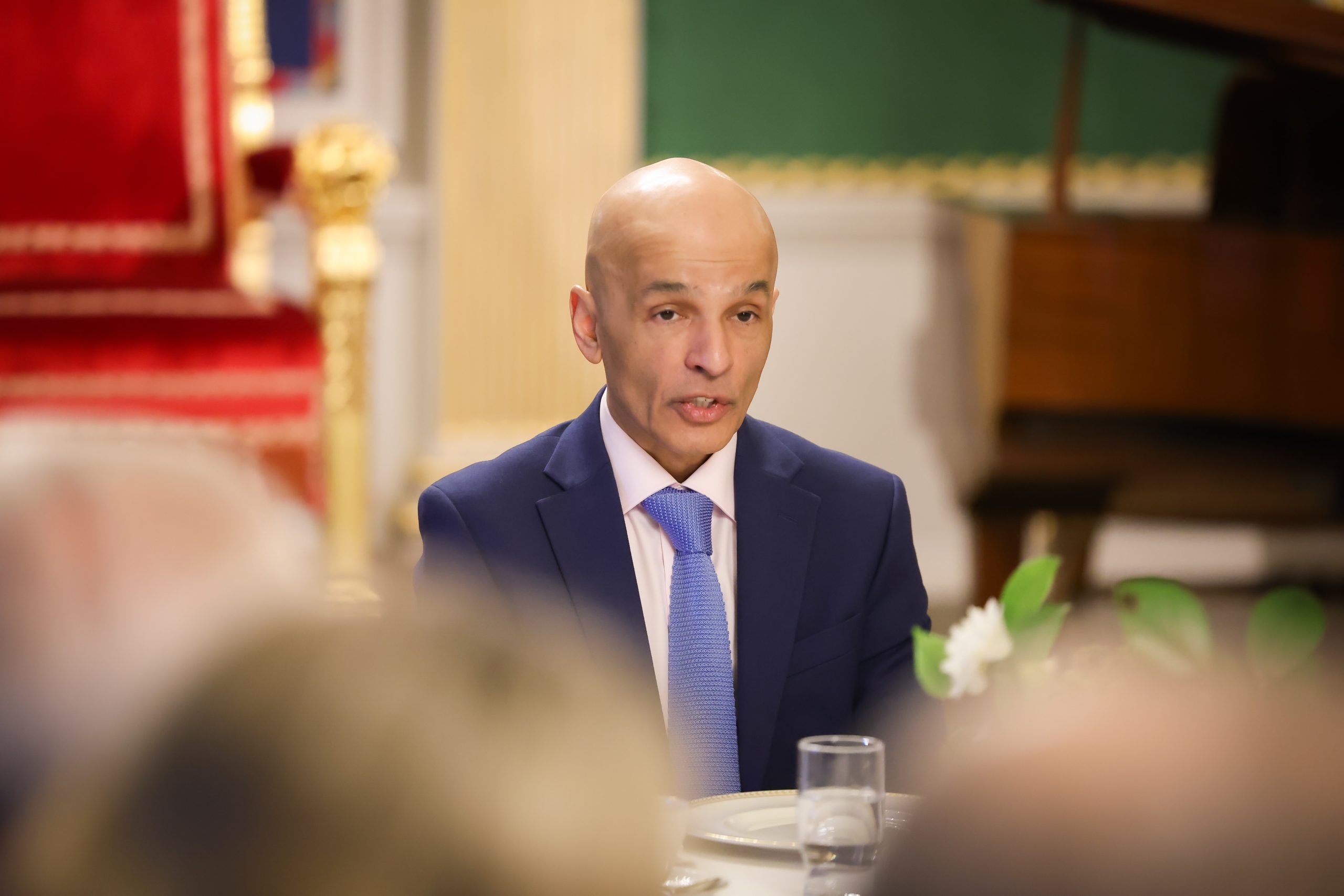 A person in formal attire seated at a table set for a meal, with a piano and ornate wall decor visible in the background.