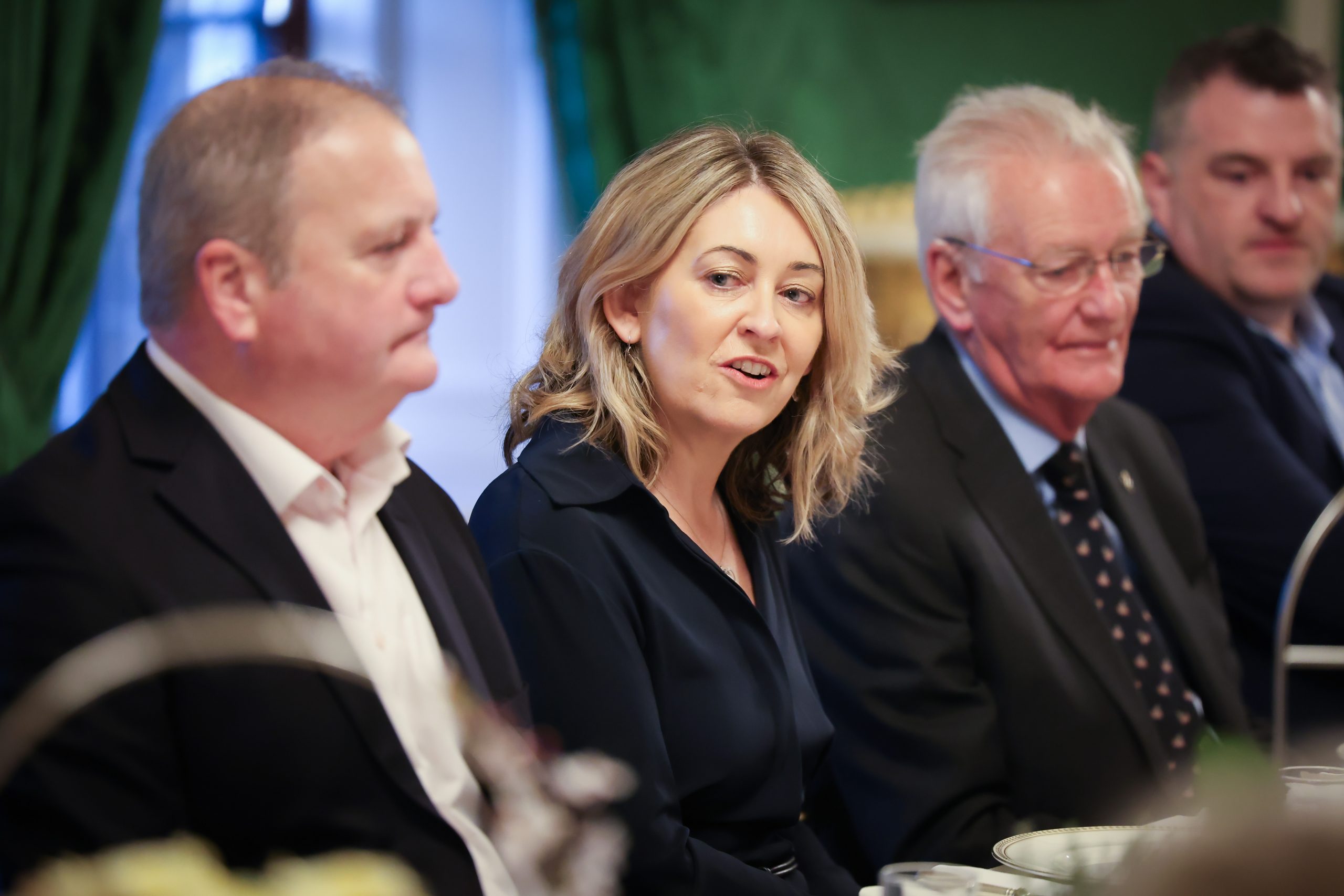 Several people seated closely together at a formal meal inside a room with green walls, framed paintings, and a window behind them.