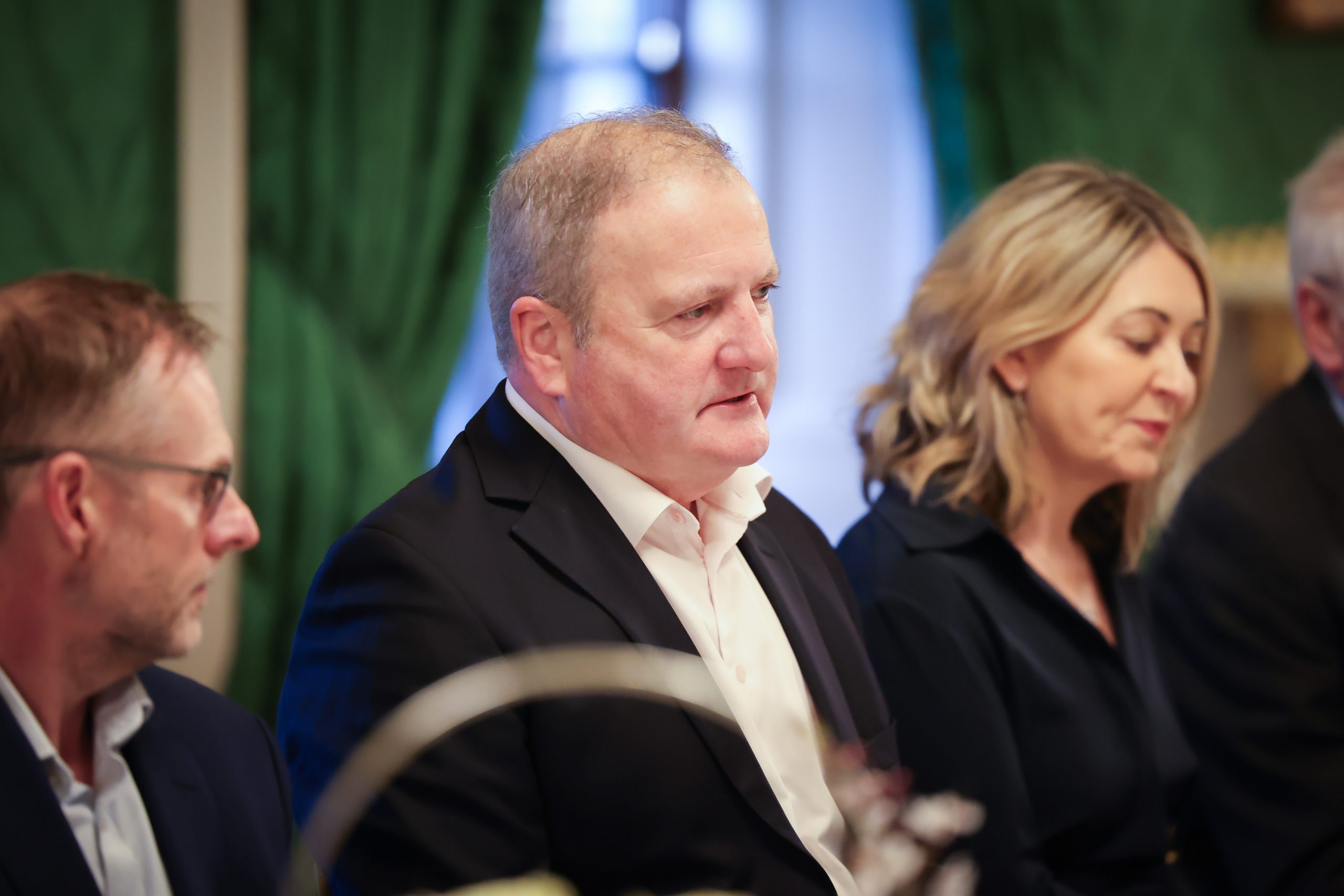 A person wearing a dark suit seated at a dining table in an ornately decorated room with green curtains and gold accents.