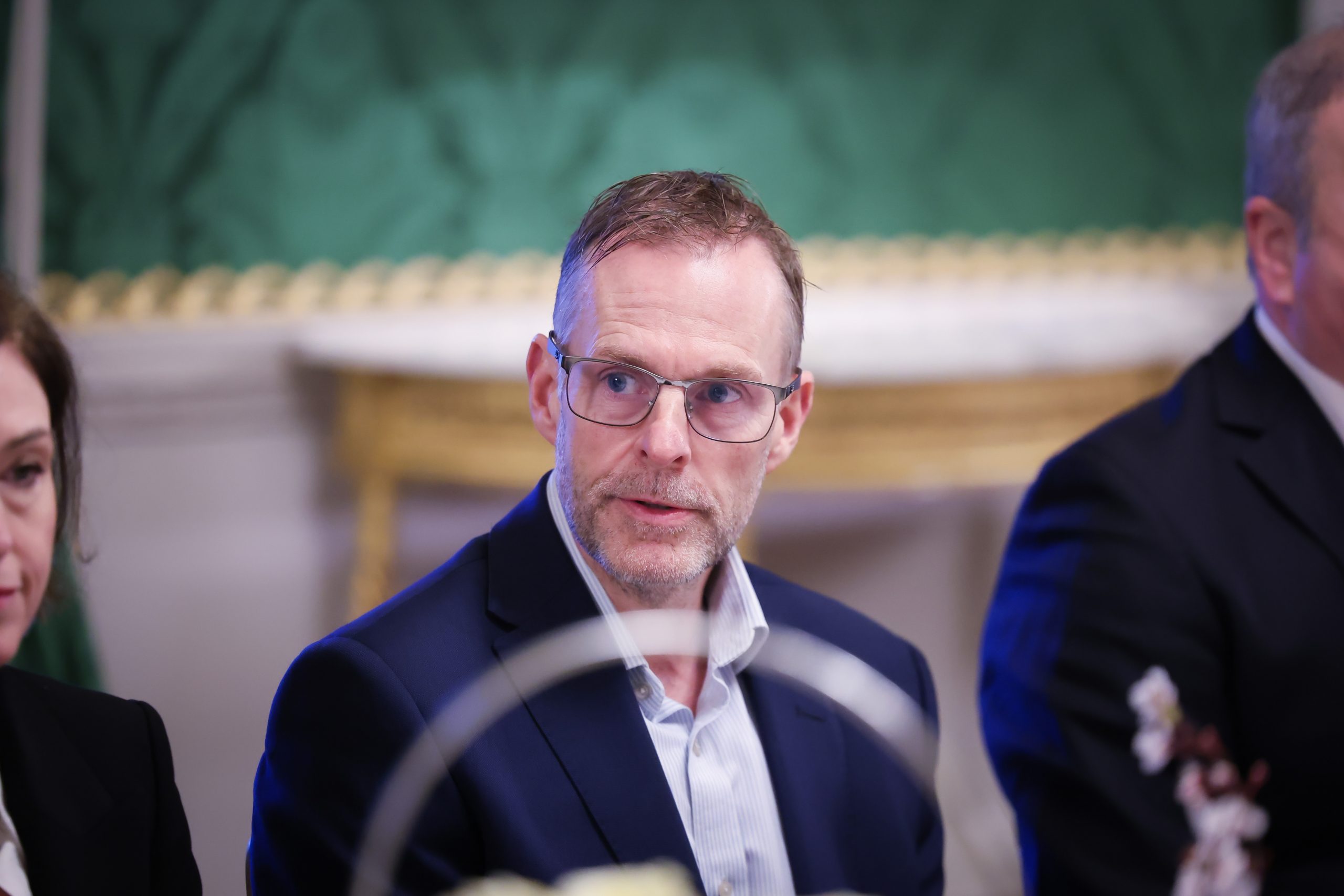 A person seated at a formal dining table in an ornate room with green walls, gold detailing, and a decorative console table in the background.