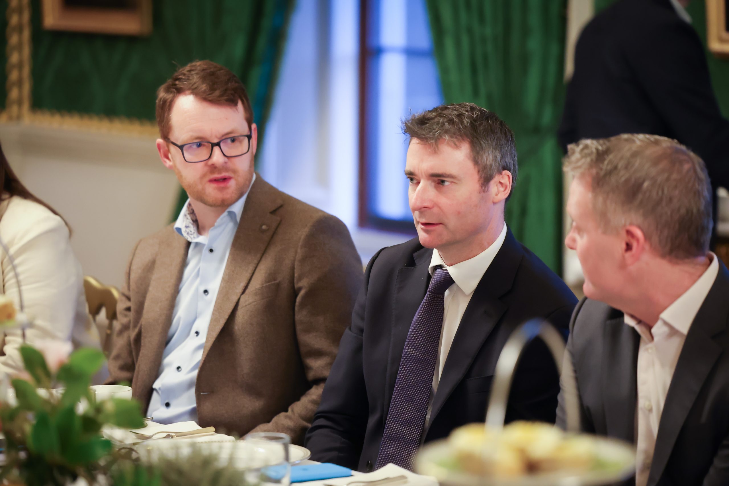 A row of attendees seated at a formal dining table, with one person wearing a brown jacket and another in a dark suit, in a room with framed artwork.