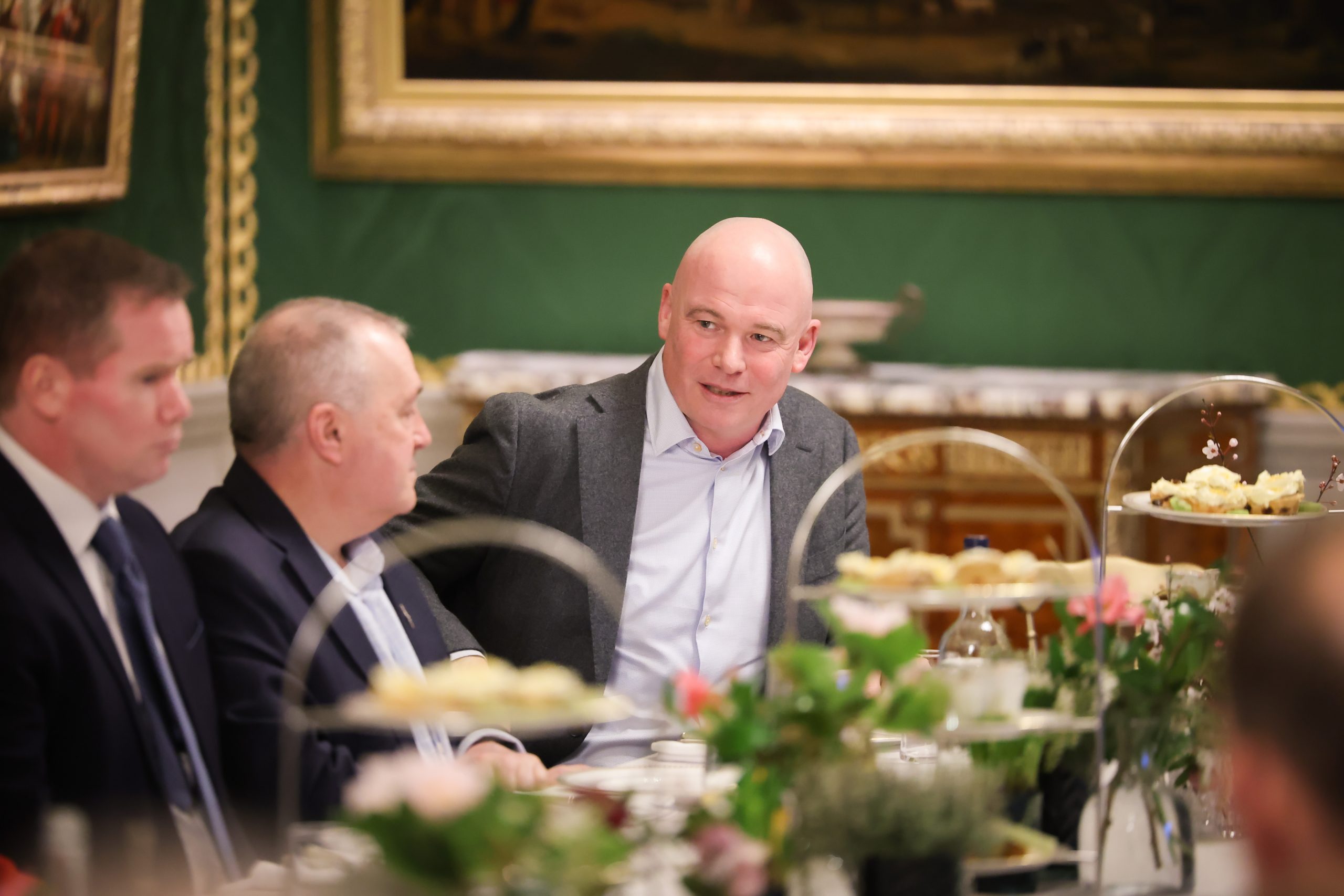 Several people seated at a formal dining table decorated with floral arrangements and tiered trays of food, in a room with ornate green walls and framed artwork.