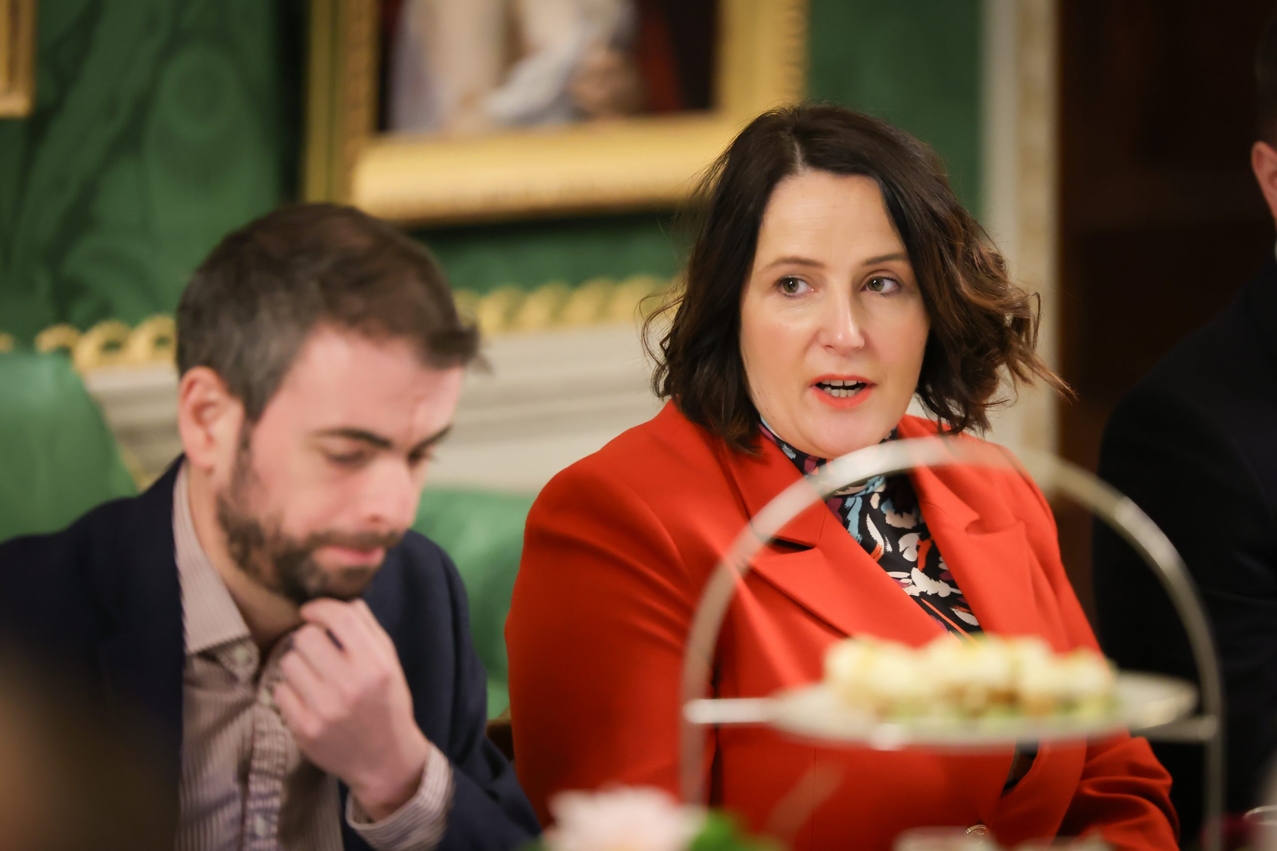 Two people seated at a formal dining table with floral arrangements and tiered trays of food, in a room decorated with framed paintings and green walls.