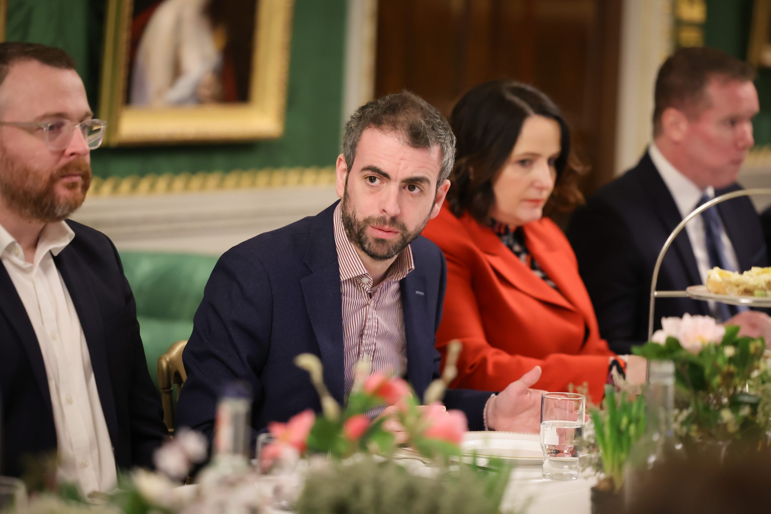 Several people seated along a dining table in an ornate room with green walls, floral centrepieces, and framed artwork.