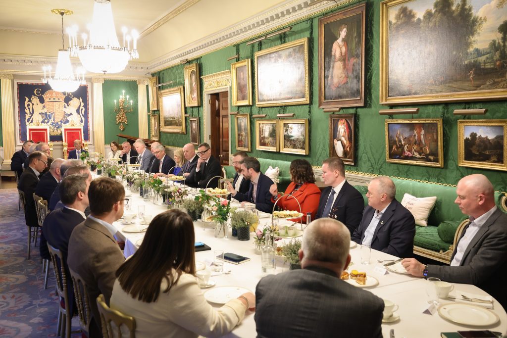 A long table of attendees seated for a formal meal inside an ornate room featuring green walls, framed artwork, and chandeliers.
