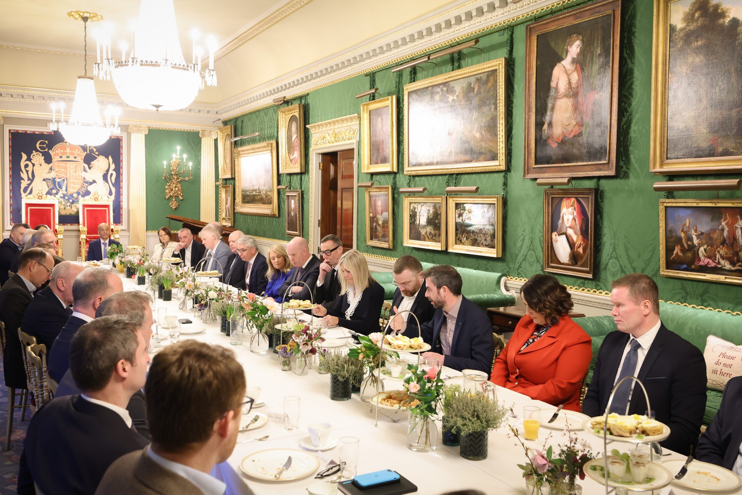 Attendees seated along a dining table set with plates, cups, floral arrangements, and food trays, with a wall of framed artwork behind them.
