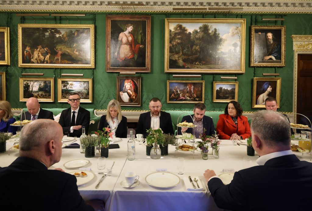  A wide shot of a formal dining table surrounded by attendees, with green walls, ornate detailing, and numerous framed paintings.