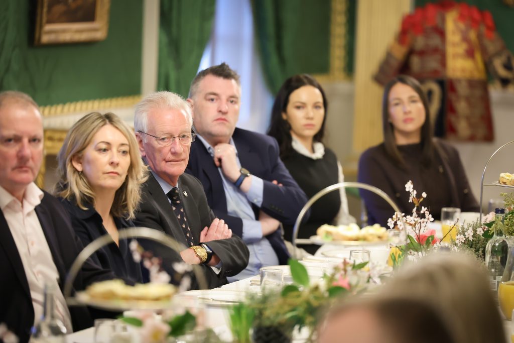 Several people seated at a long dining table with food trays, floral centrepieces, and green wall décor in the background.