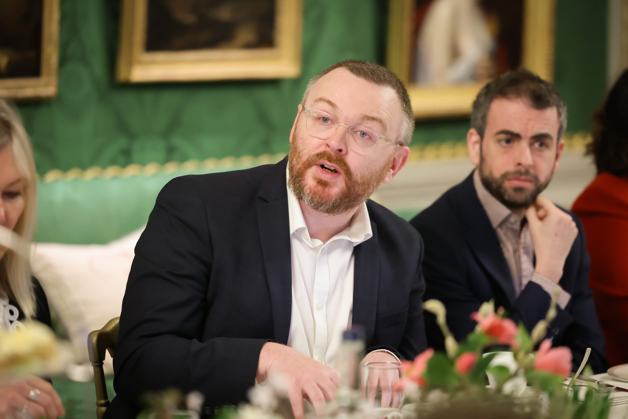 person seated at a dining table leaning slightly forward while speaking. Framed artwork and table flowers decorate the room.