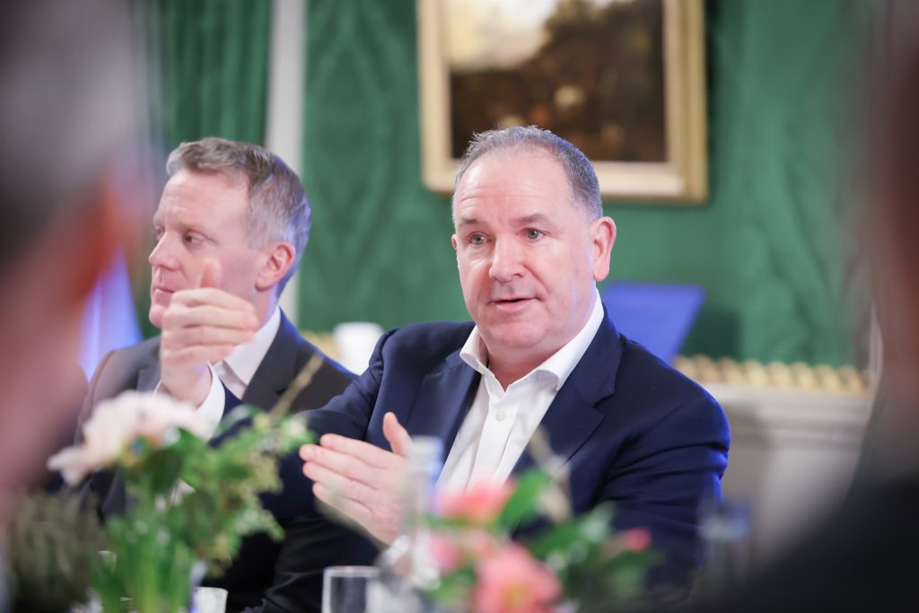  Two people seated beside each other at a dining table. One has hands raised mid‑gesture. Green drapes and a framed landscape painting decorate the wall behind.