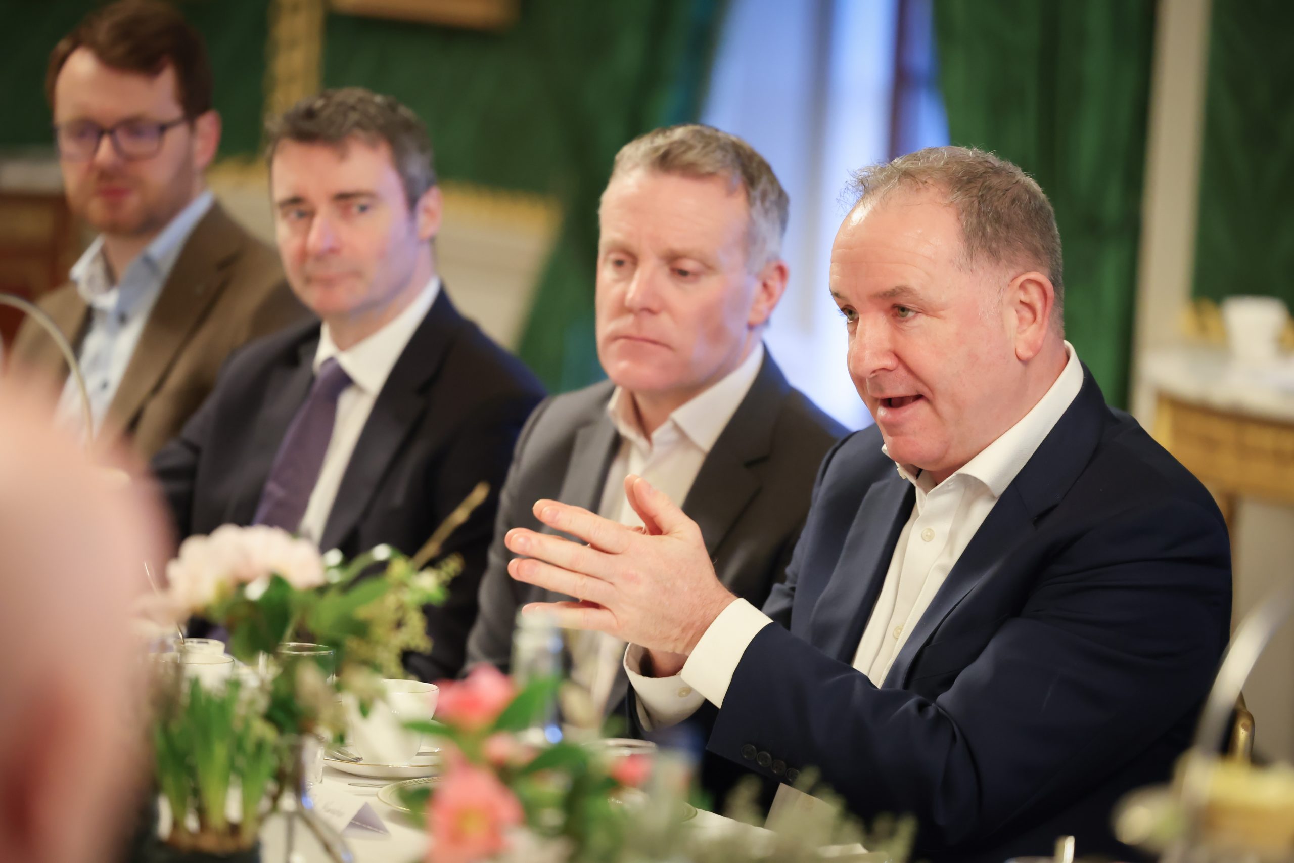 Three people seated at a formal dining table, with one person speaking and another writing in a notebook. Floral arrangements line the centre of the table.