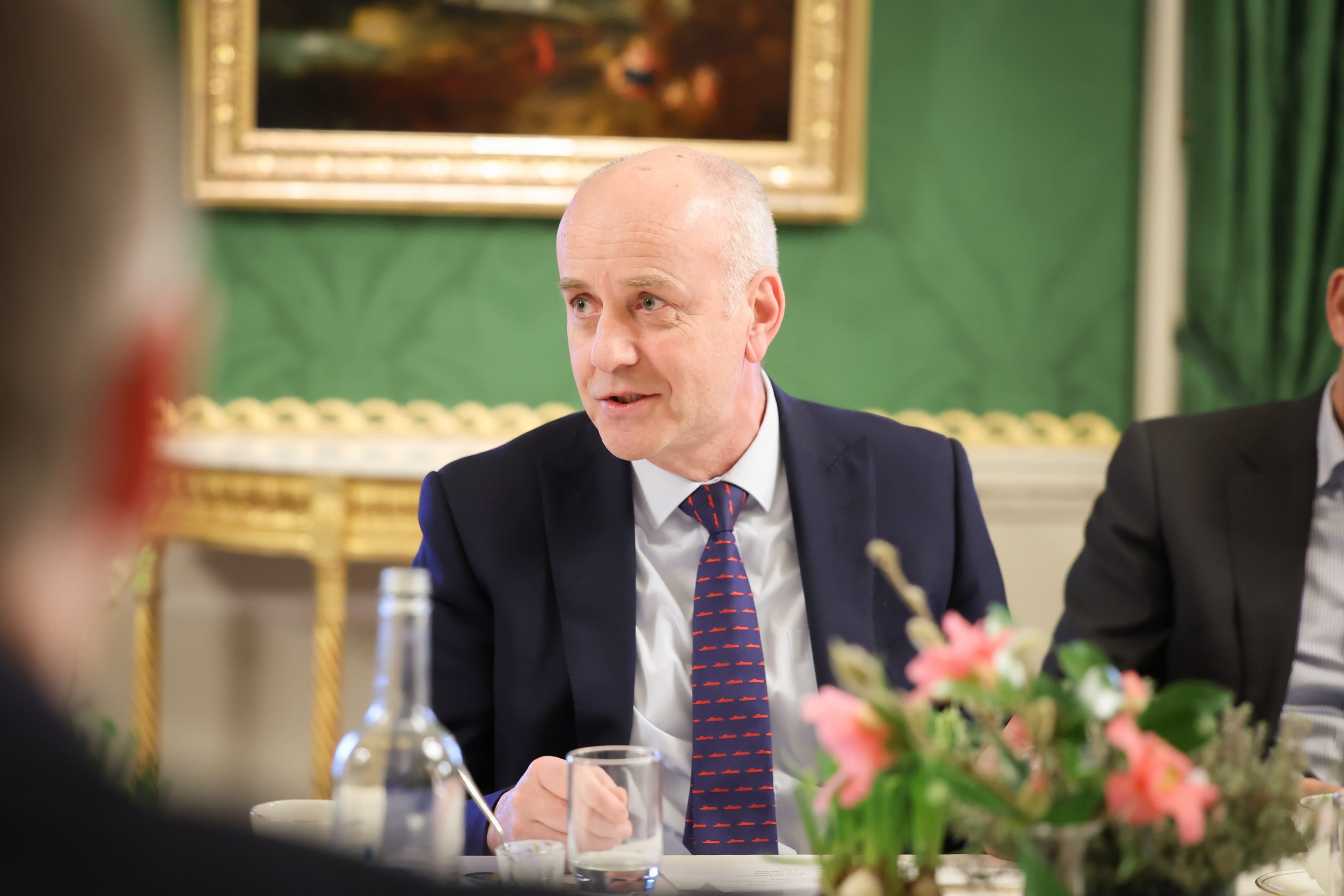 A person seated at a long dining table with a water bottle and glass in front of them, surrounded by flowers and ornate decor.